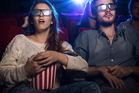 young teenagers at the cinema wearing glasses and watching a 3d movie, a girl is eating popcorn, entertainment and movies concept
