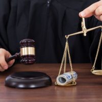 midsection of male judge striking gavel while holding scale with money in courtroom