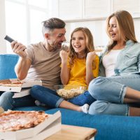 Photo of excited beautiful caucasian family eating popcorn and pizza while watching tv at home