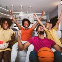 Happy African American family of three watching tv and cheering basketball games on sofa at home.