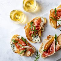 Appetizer crostini, tapas, open faced sandwiches with pear, prosciutto, arugula and blue cheese on white marble board. Delicious snack, appetizers